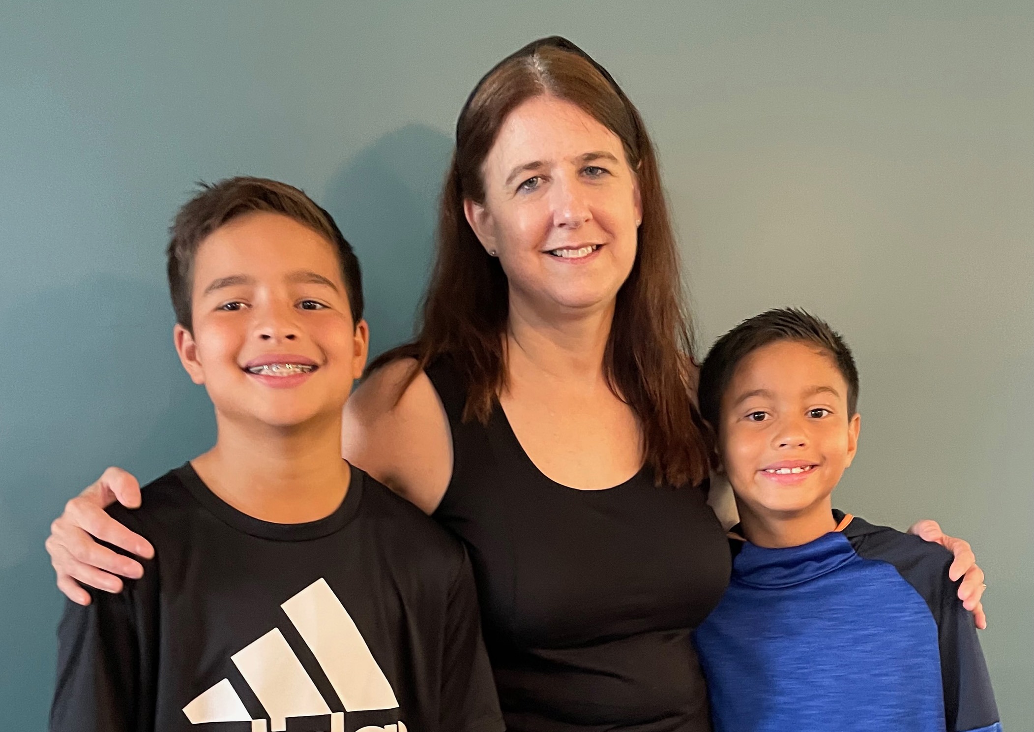 A woman smiles while standing between two boys, one in a black shirt and the other in a blue shirt, against a teal background.