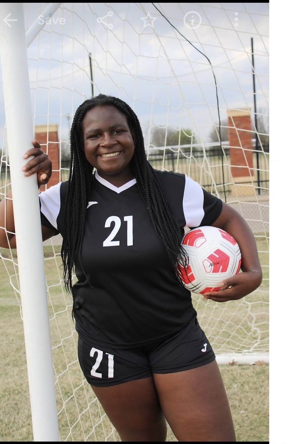 Young female soccer player in black uniform, number 21, smiling confidently while posing with a soccer ball by the goalpost on a field.