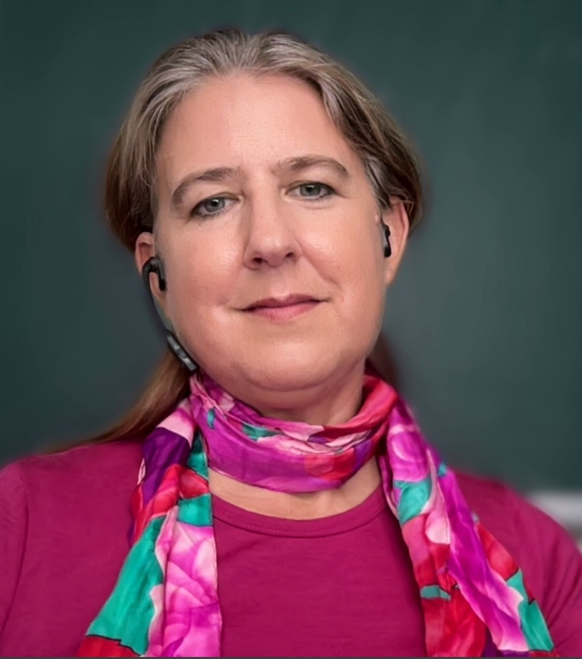 A woman with long, reddish-brown hair wears a colorful floral scarf and wireless earbuds, smiling confidently against a neutral background.