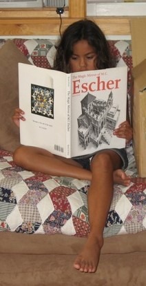 A child sits on a sofa, engrossed in a book about M.C. Escher, surrounded by a colorful quilt. The image highlights curiosity and appreciation for art.
