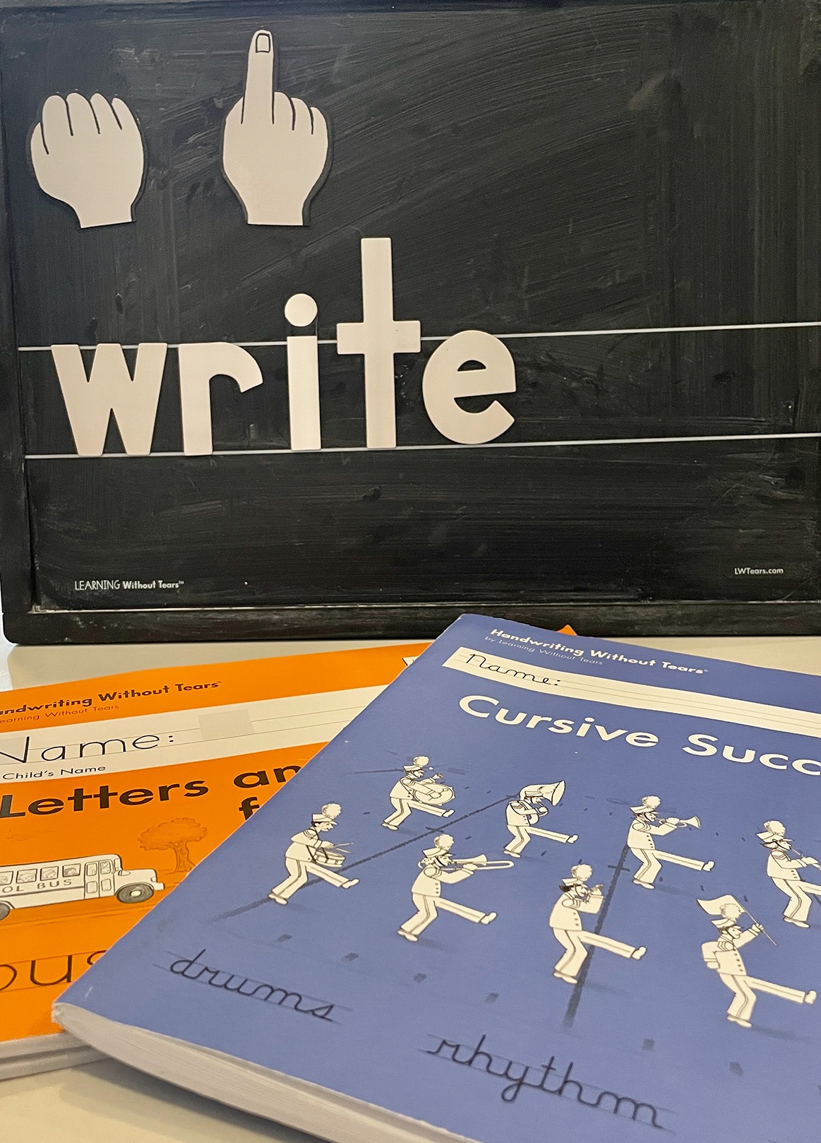 Blackboard displaying "write" alongside hand symbols, with handwriting workbooks titled "Letters and Numbers" and "Cursive Success" prominently featured.