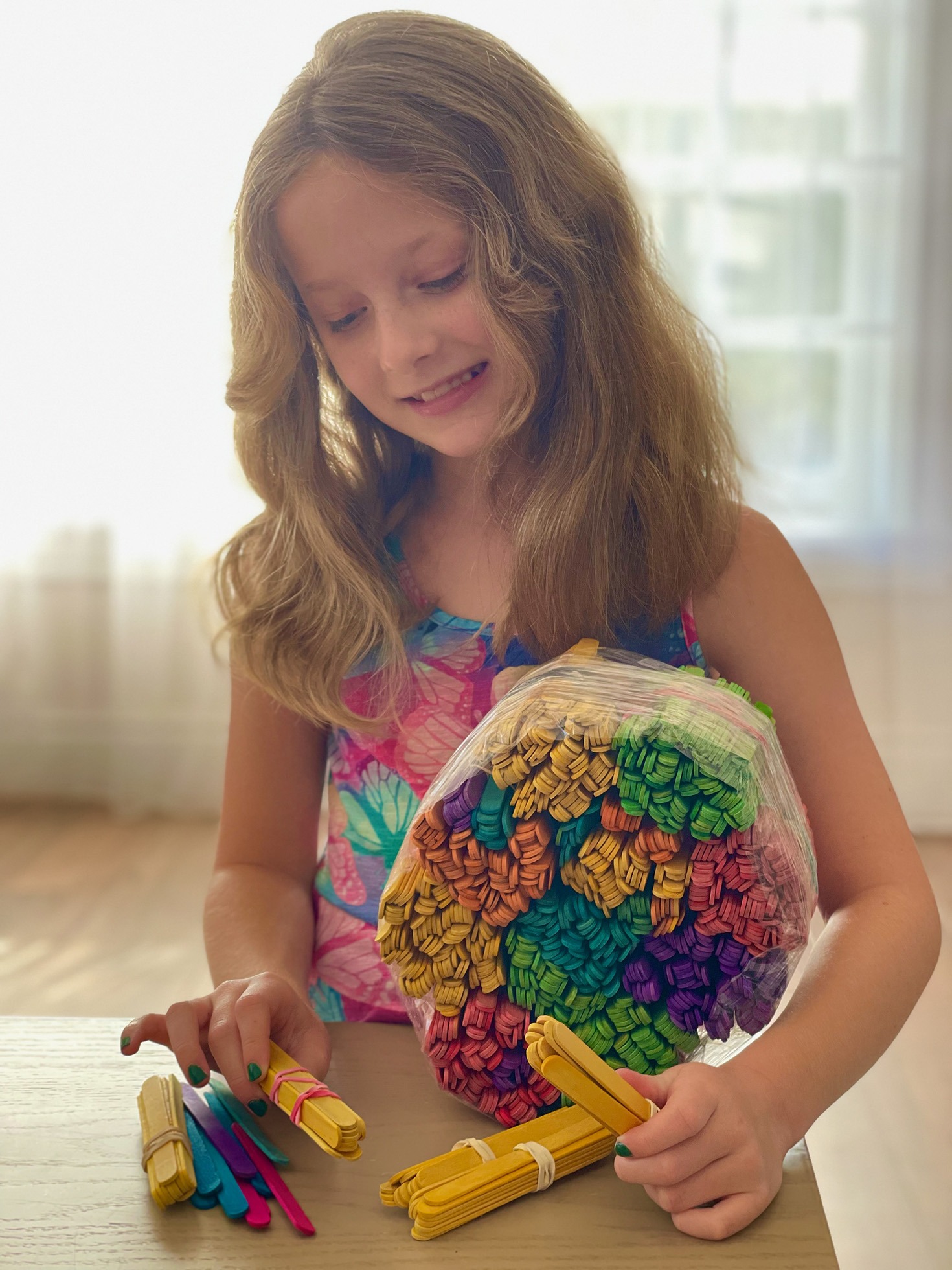 A smiling girl with wavy hair sorts colorful craft sticks on a table, showcasing her creativity and engaging with art supplies.