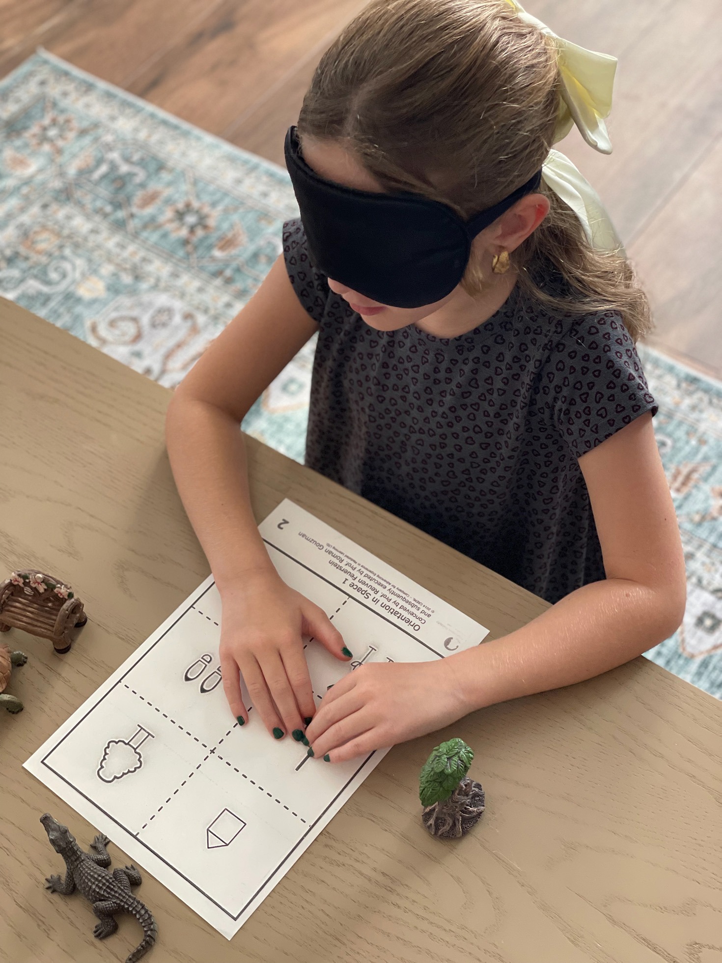 Child wearing a black eye mask engages in a tactile learning activity, feeling different objects around a worksheet with shapes.