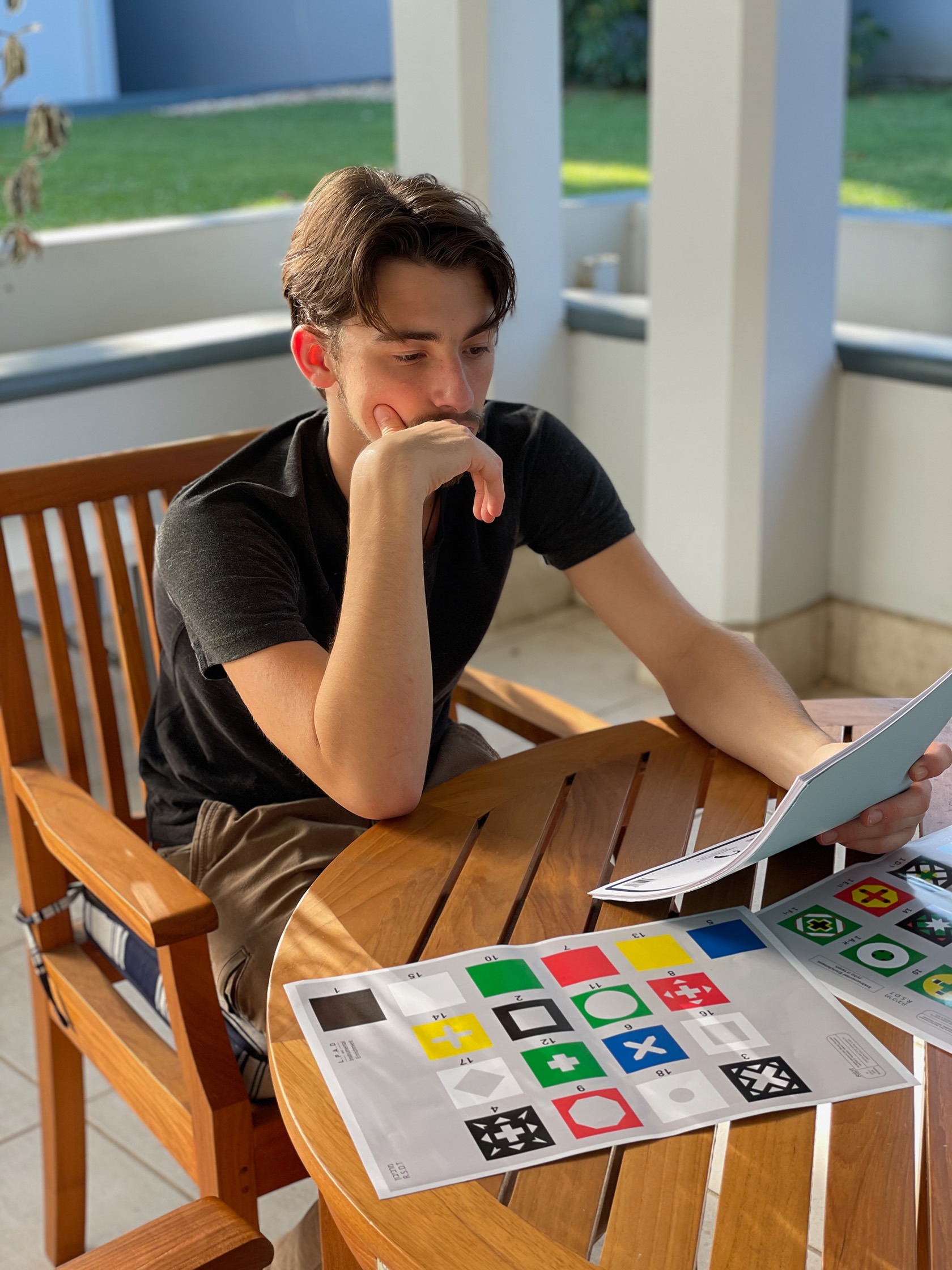 A young man sits at a wooden table reviewing colored flag designs laid out in front of him, deep in thought.