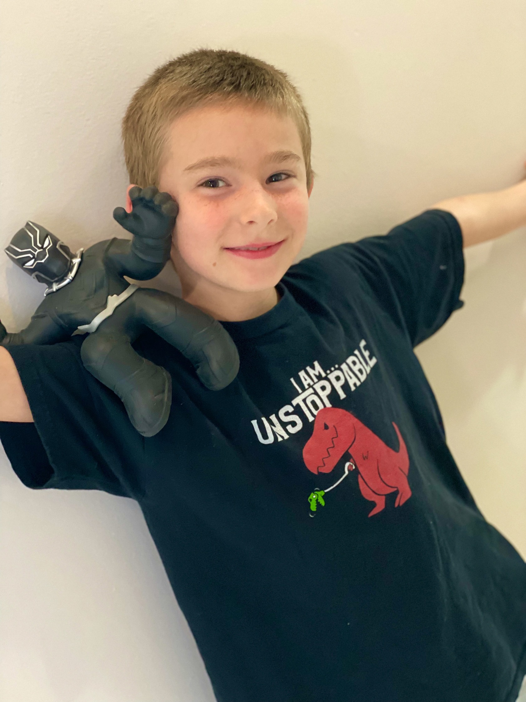 Young boy with short hair smiles while holding a Black Panther action figure, wearing a t-shirt that reads "I AM UNSTOPPABLE," against a white wall.