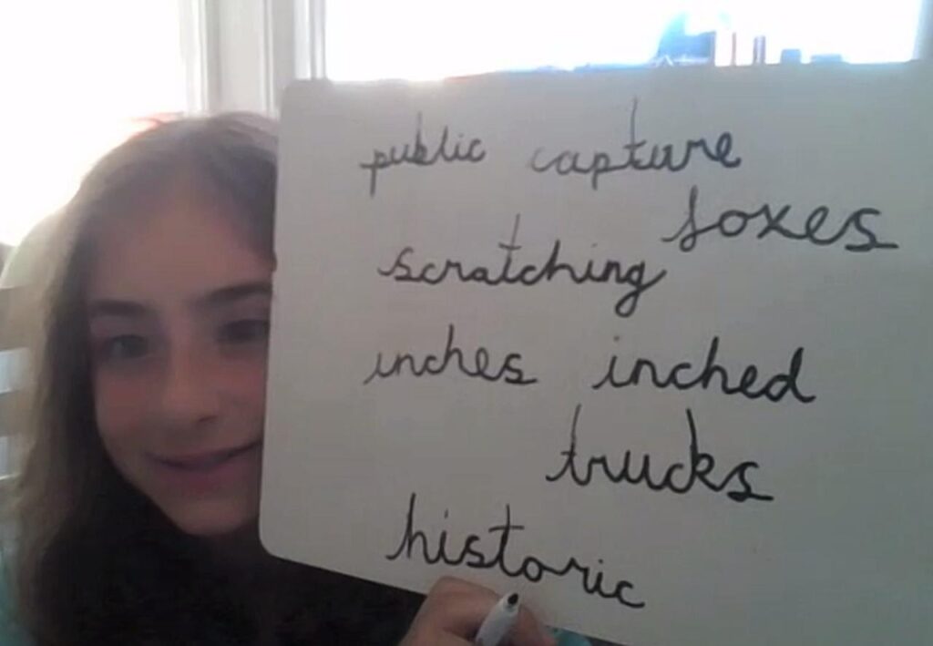 A young girl holds a whiteboard displaying a list of words: "public," "capture," "scratching," "boxes," "inches," "inched," "trucks," and "historic."
