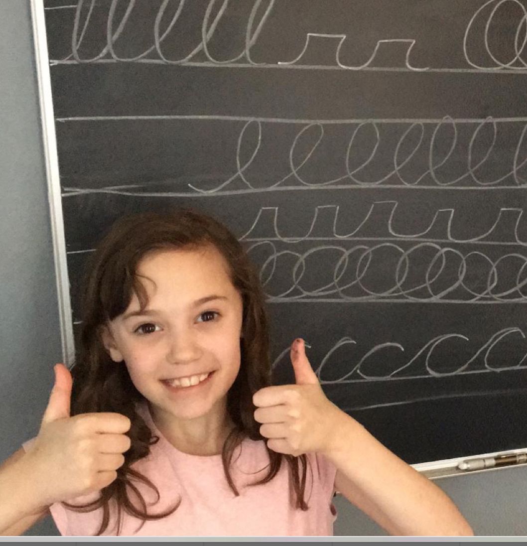 A smiling girl gives two thumbs up in front of a chalkboard with writing practice lines, showcasing her enthusiasm for learning.